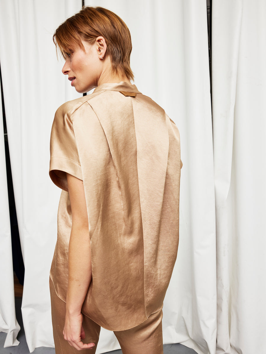 A person with short brown hair stands with their back to the camera in front of white curtains, wearing the Fin Gaban Shirt and matching pants by Zero + Maria Cornejo in shiny gold satin with a stand collar.