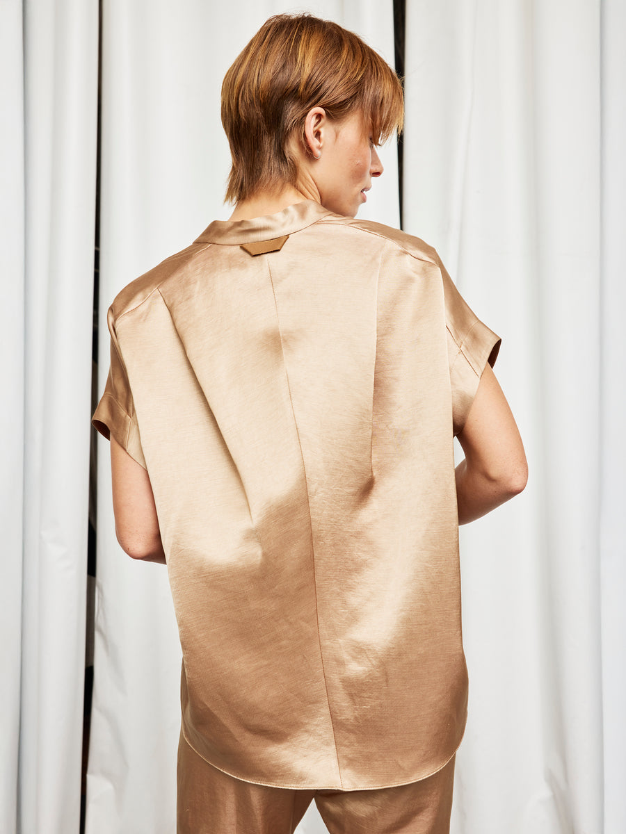 A person with short, light brown hair stands with their back to the camera, wearing the shiny beige Fin Gaban Shirt by Zero + Maria Cornejo and matching pants, in front of white curtains.