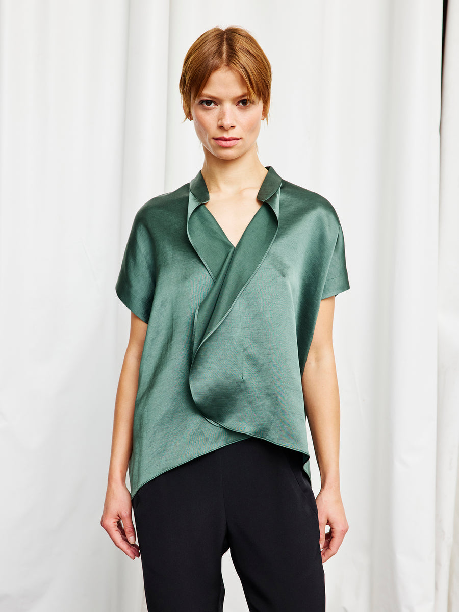A person with short light brown hair wears the shiny green Fin Gaban Shirt by Zero + Maria Cornejo, styled with ruffle detail and black pants, standing in front of white curtains.