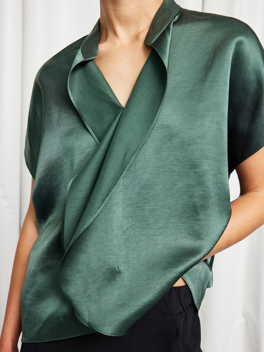 A person wears the Fin Gaban Shirt by Zero + Maria Cornejo—a shiny, dark green silk blouse with ruffle details and a wide, overlapping collar—paired with black pants in front of a plain white curtain.