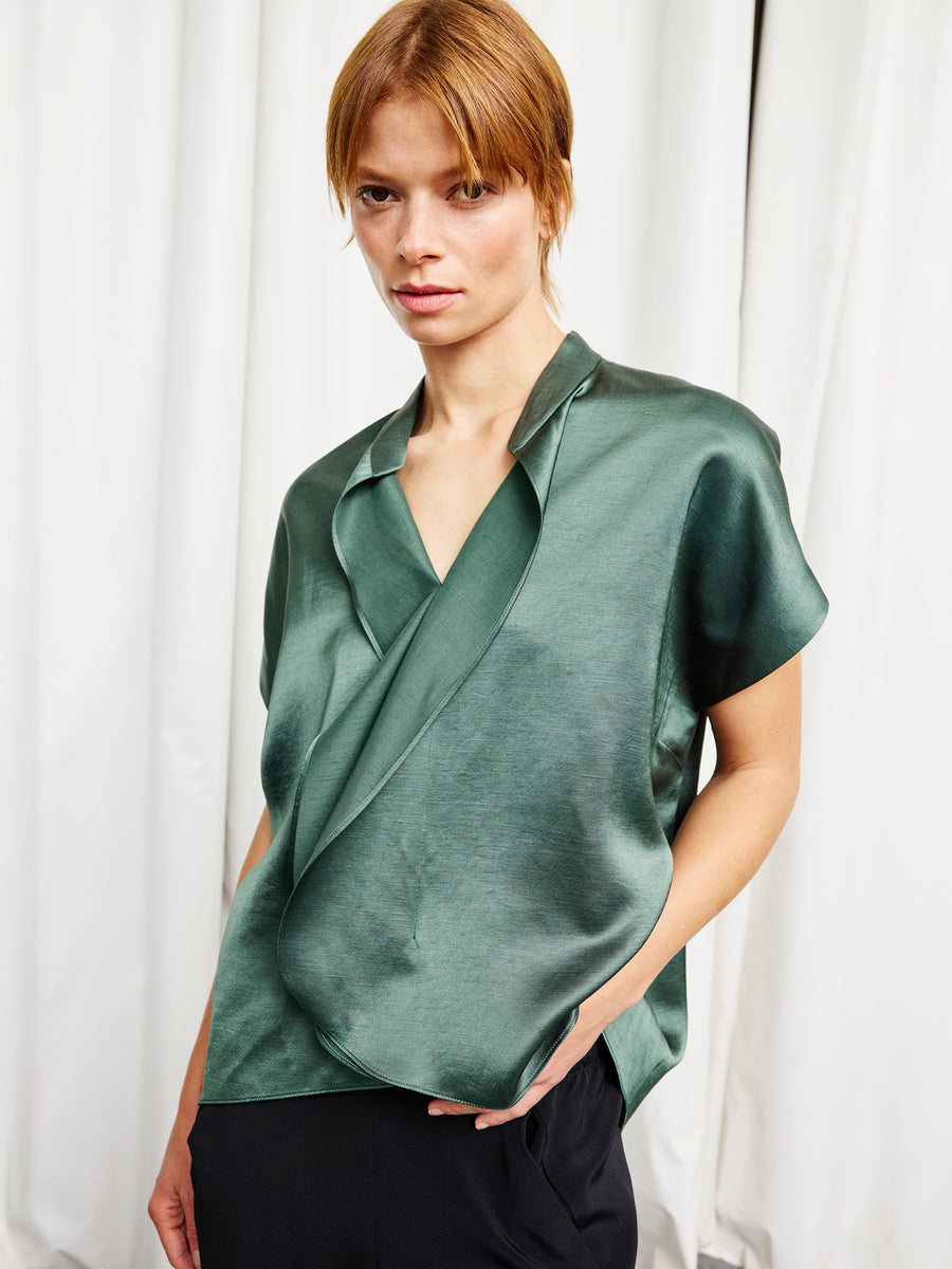 A woman with short, light brown hair wears the shiny green satin Fin Gaban Shirt by Zero + Maria Cornejo with black pants, standing in front of white curtains.