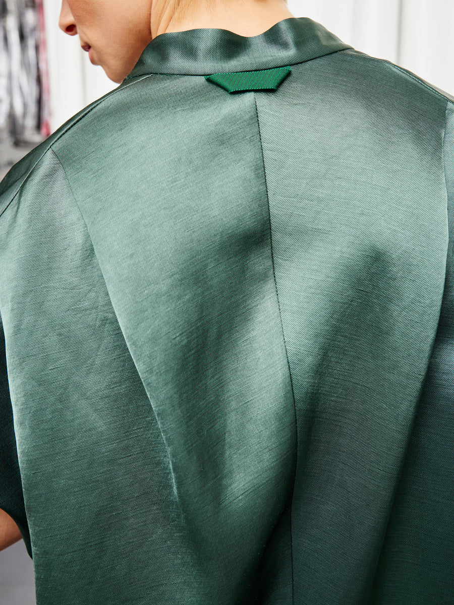 Close-up of a person in the Fin Gaban Shirt by Zero + Maria Cornejo, featuring shiny green linen-blend fabric, smooth texture, center seam, and a small green tab near the collar. The face is partially visible on the left.