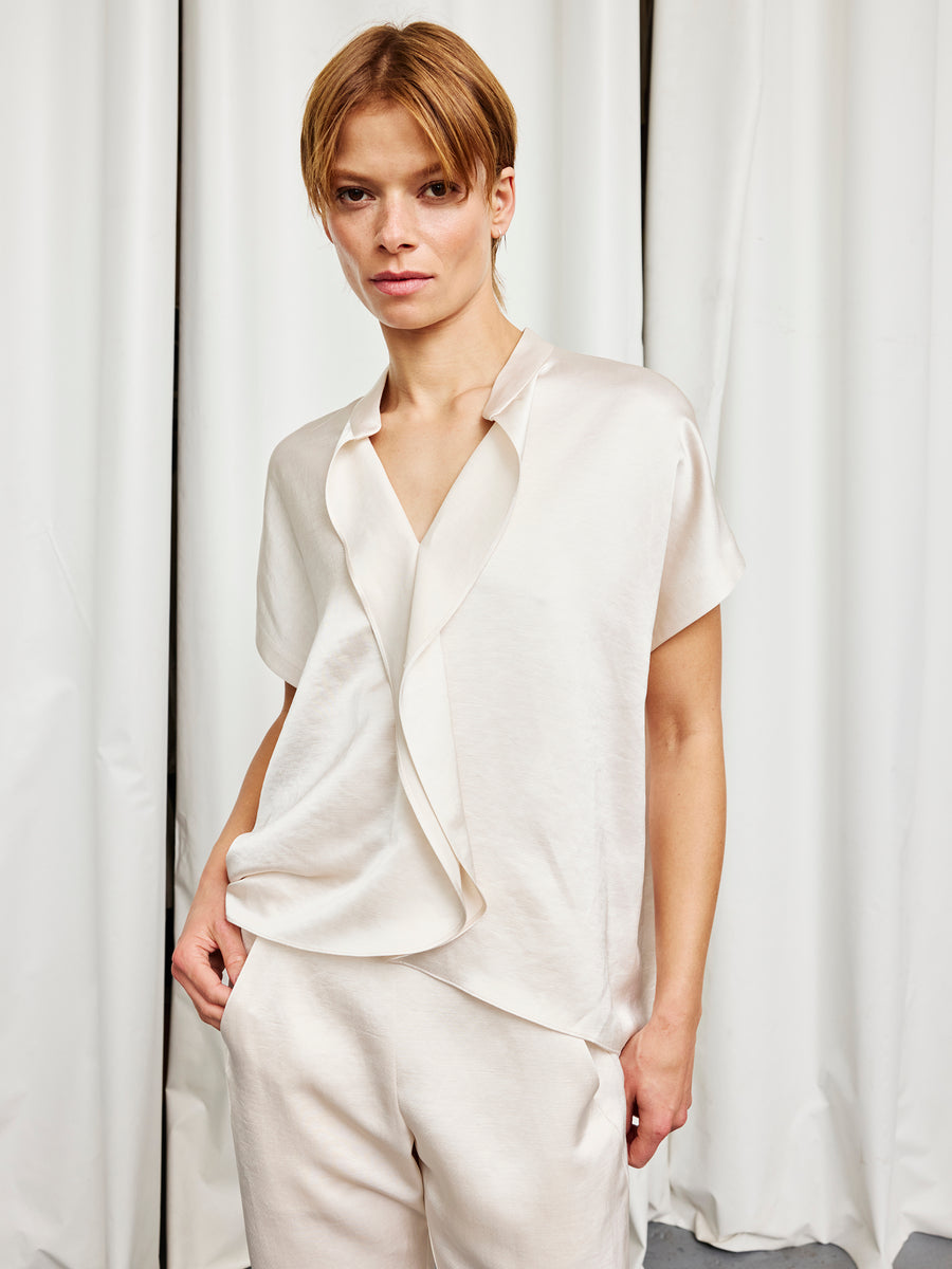 A person with short light brown hair wears the cream-colored Fin Gaban Shirt by Zero + Maria Cornejo, paired with matching pants, standing before white curtains with one hand in their pocket.