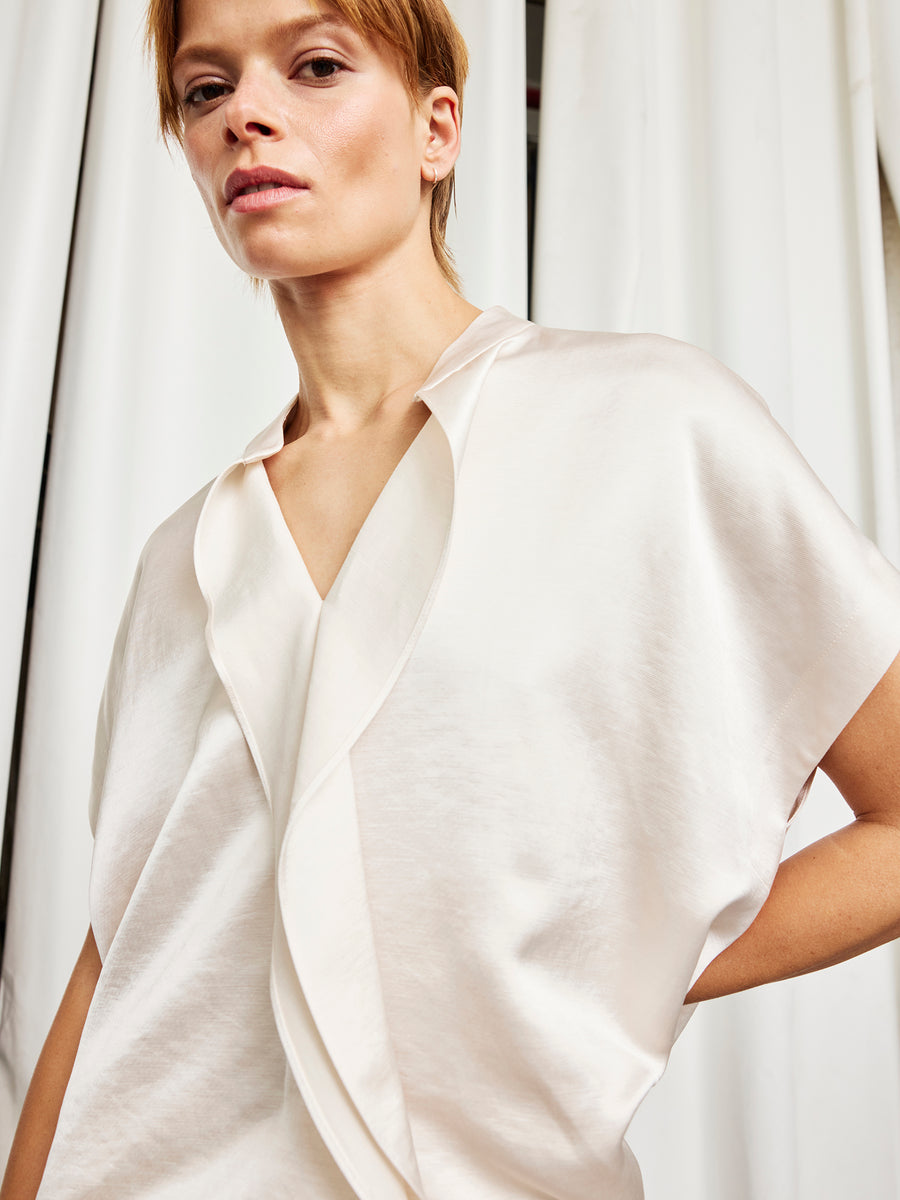 A person with short light brown hair wears the Zero + Maria Cornejo Fin Gaban Shirt in Porcelain, a loose, shiny blouse with a stand collar and deep V-neck, standing before white curtains and gazing slightly upward.