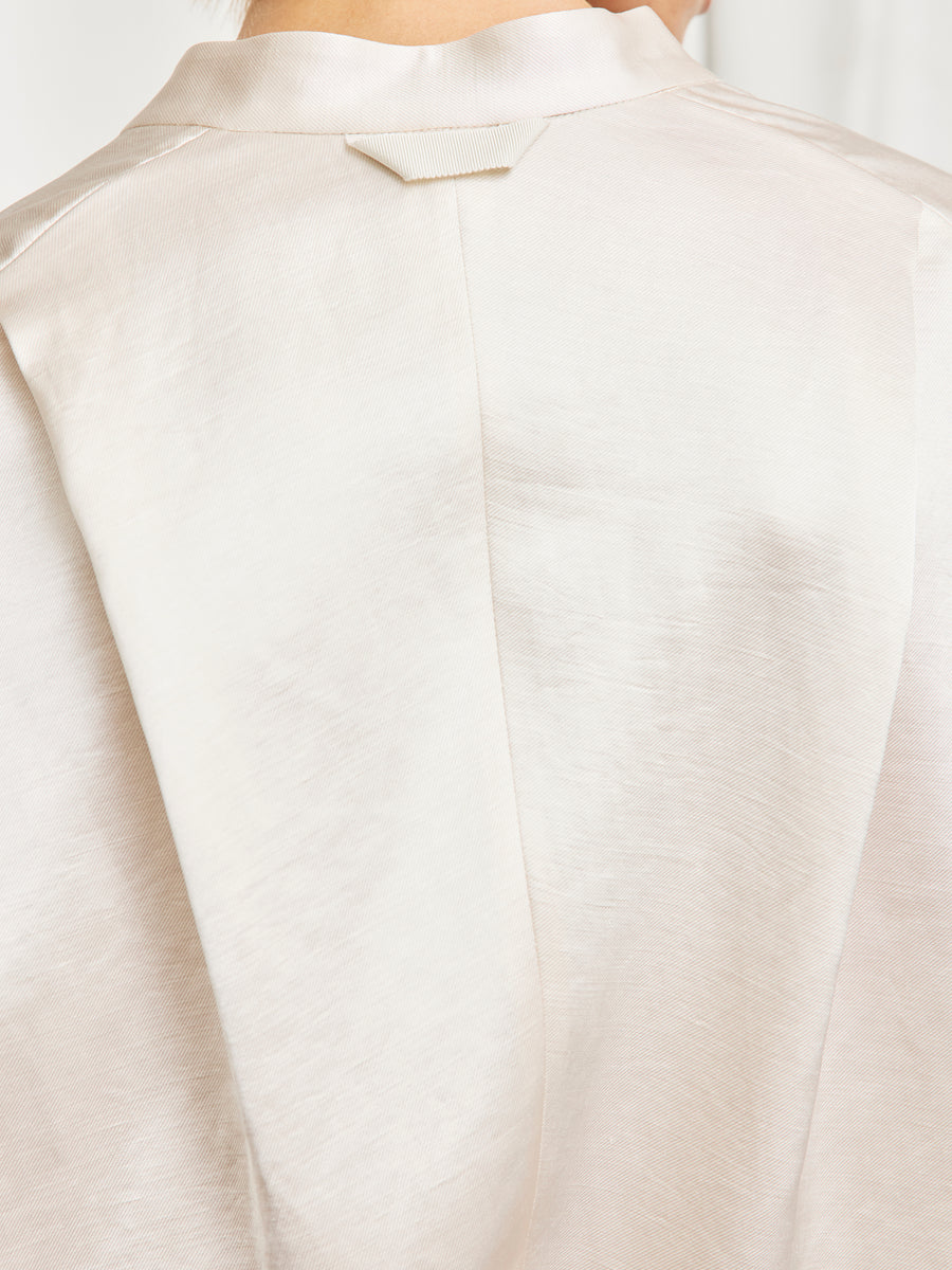 Close-up of the upper back of a person wearing the Fin Gaban Shirt by Zero + Maria Cornejo, showing its light, silky cream-colored linen-blend fabric and a small rectangular loop detail near the stand collar.