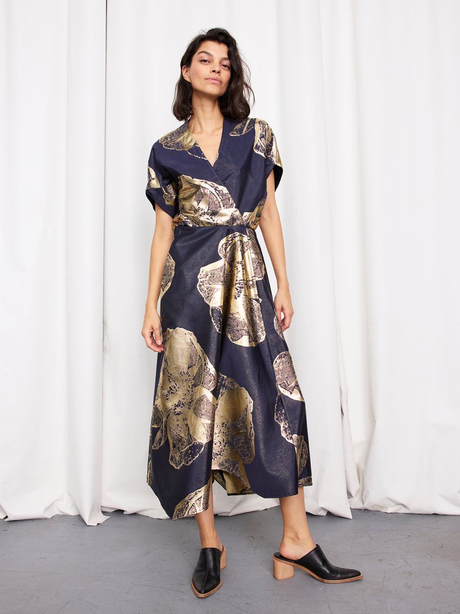 A woman with dark hair stands before white curtains in the Zero + Maria Cornejo Long Aki Wave Dress, a dark blue floral jacquard midi with gold accents, a tied waist, and short sleeves, paired with black mule heels featuring wooden block heels.