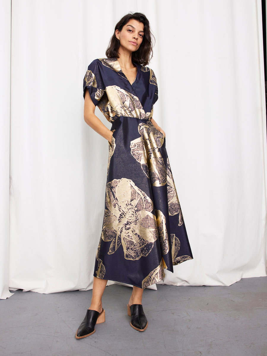A woman poses confidently in the Zero + Maria Cornejo Long Aki Wave Dress, featuring a navy blue mid-length design with gold leaf patterns, short sleeves, a V-neck, pockets, and paired with black mule shoes in front of white curtains.