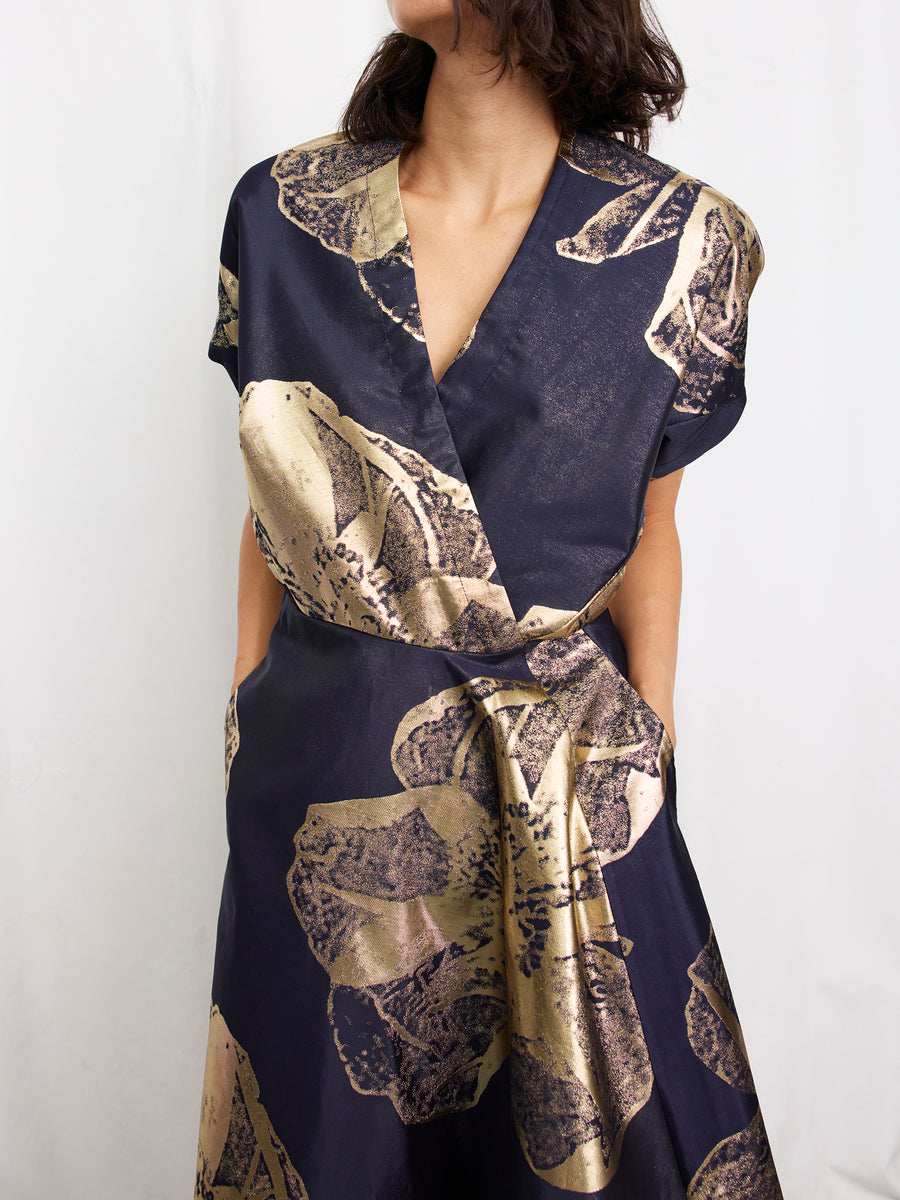 A woman stands against a white background, hands in her pockets and head slightly turned, wearing the Zero + Maria Cornejo Long Aki Wave Dress—navy blue with a shiny gold abstract leaf pattern.