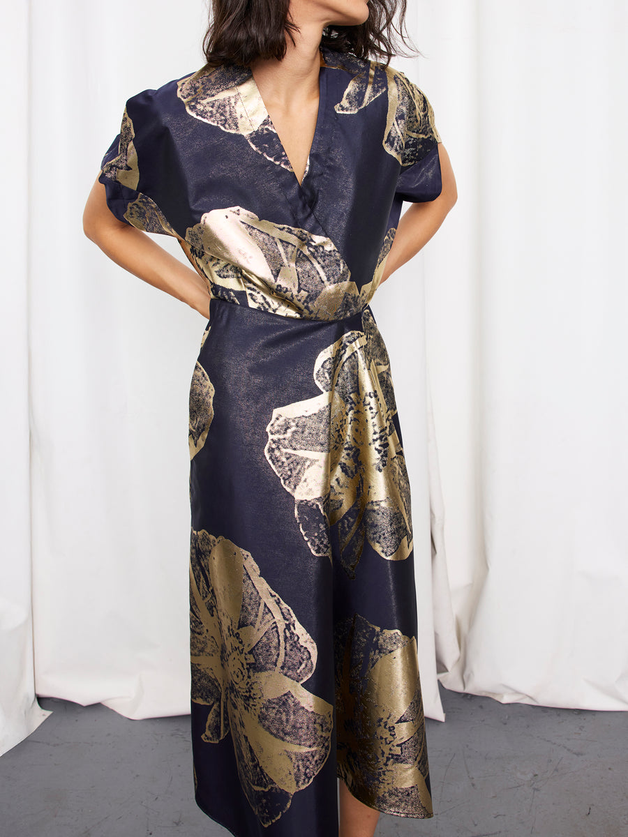 A woman stands with her hands on her hips in front of white curtains, wearing the Zero + Maria Cornejo Long Aki Wave Dress, a navy mid-length piece with gold floral jacquard, short sleeves, and a deep V neckline.