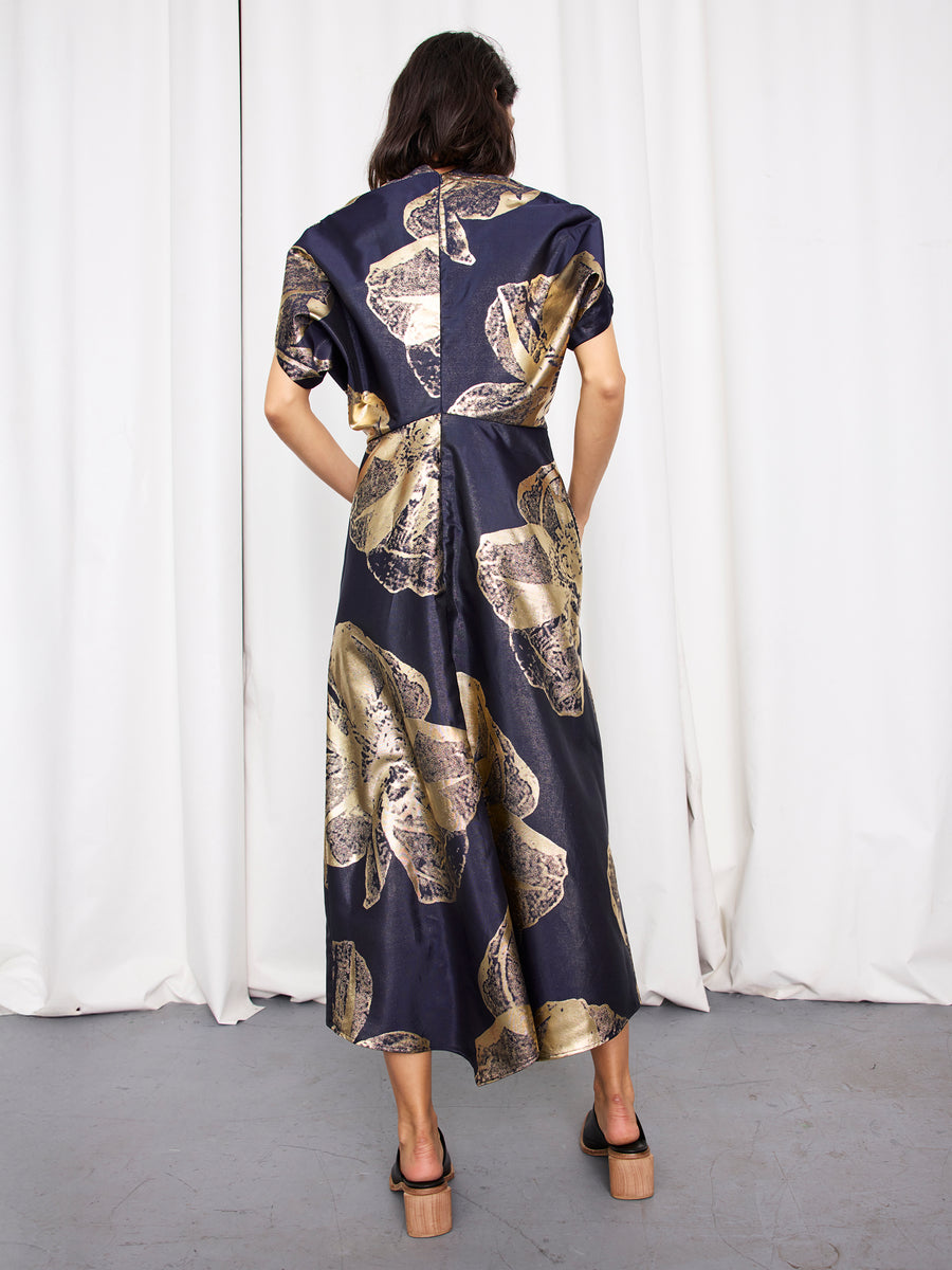 A person faces away, wearing the Zero + Maria Cornejo Long Aki Wave Dress—dark blue with a gold floral jacquard pattern and short sleeves—paired with black heeled sandals. White curtains and a gray floor form an elegant backdrop.