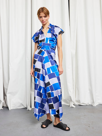 A person stands before white curtains wearing the Long Ruched Stella Dress by Zero + Maria Cornejo—a full-length, blue, white, and black geometric patterned, short-sleeve dress paired with black slide sandals.