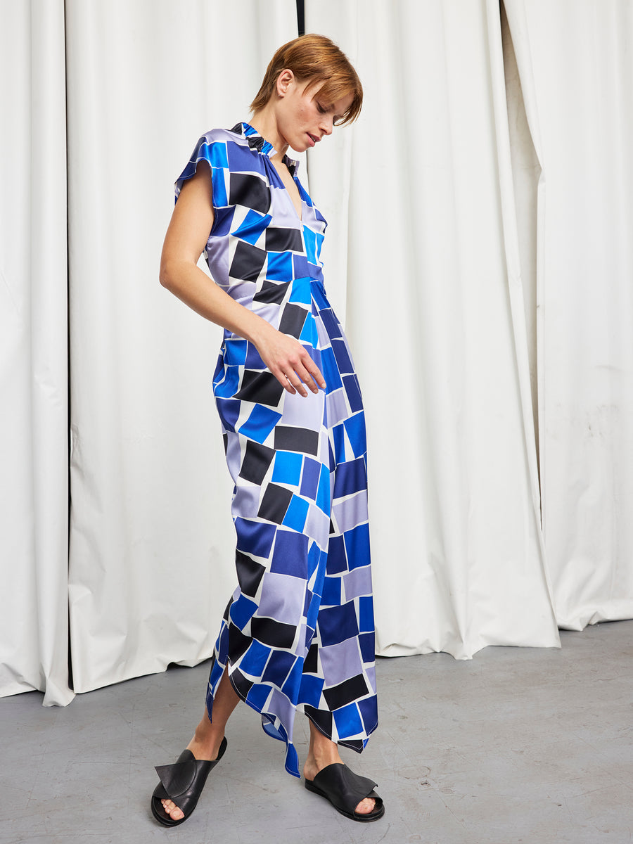 Wearing the Long Ruched Stella Dress by Zero + Maria Cornejo—a short-sleeved, full-length silk dress with bold blue, black, and white geometric prints—a person in black sandals stands before white curtains, gazing down.