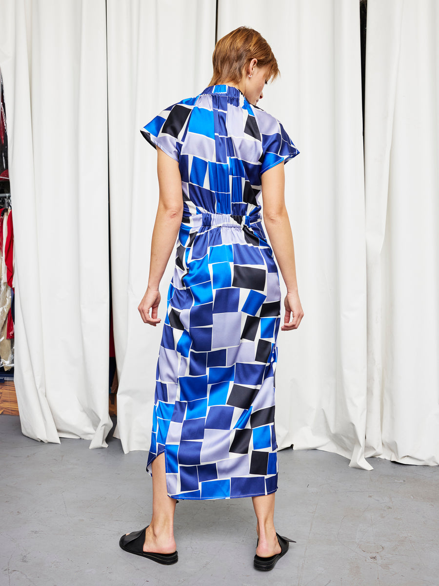 A person stands with their back to the camera in front of white curtains, wearing the Zero + Maria Cornejo Long Ruched Stella Dress in a blue, black, and white geometric pattern, paired with black slide sandals.