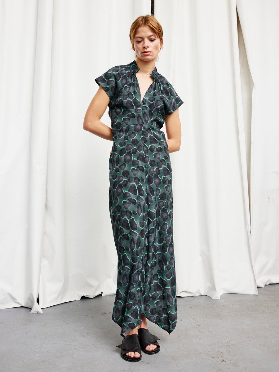 A woman with light hair pulled back stands before white curtains, wearing the Long Ruched Stella Dress by Zero + Maria Cornejo and black open-toe sandals, her arms loosely crossed.