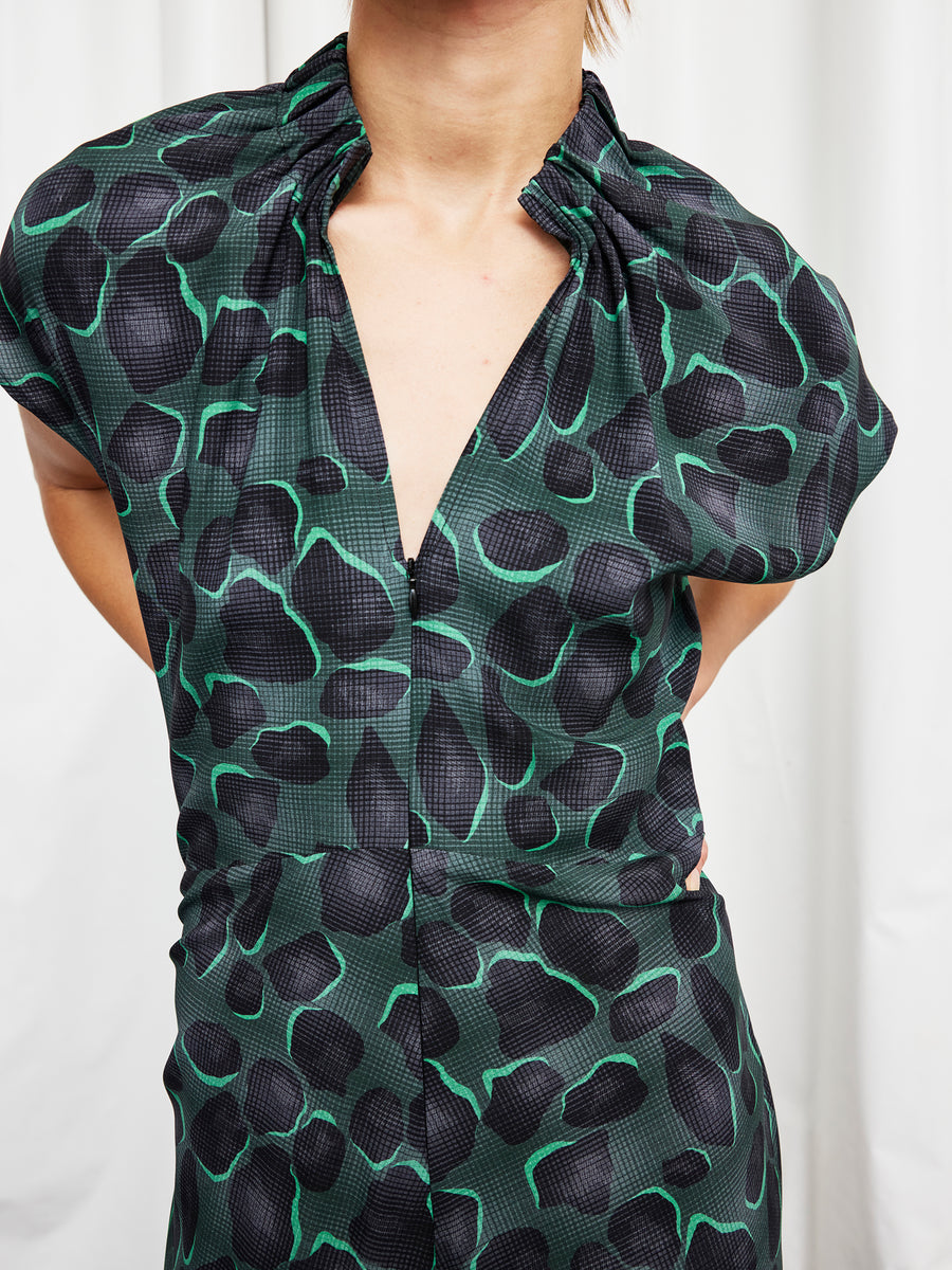 A person wearing the Long Ruched Stella Dress by Zero + Maria Cornejo—a full-length green and black animal-print silk charmeuse dress with a deep V-neck and gathered collar—stands in front of a white curtain, arms behind their back.