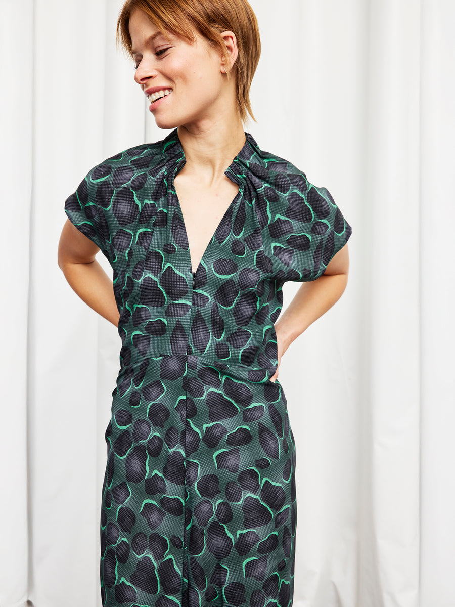 A person smiling and looking to the side, wearing the Zero + Maria Cornejo Long Ruched Stella Dress in dark green and black silk charmeuse with short sleeves, standing in front of white curtains.