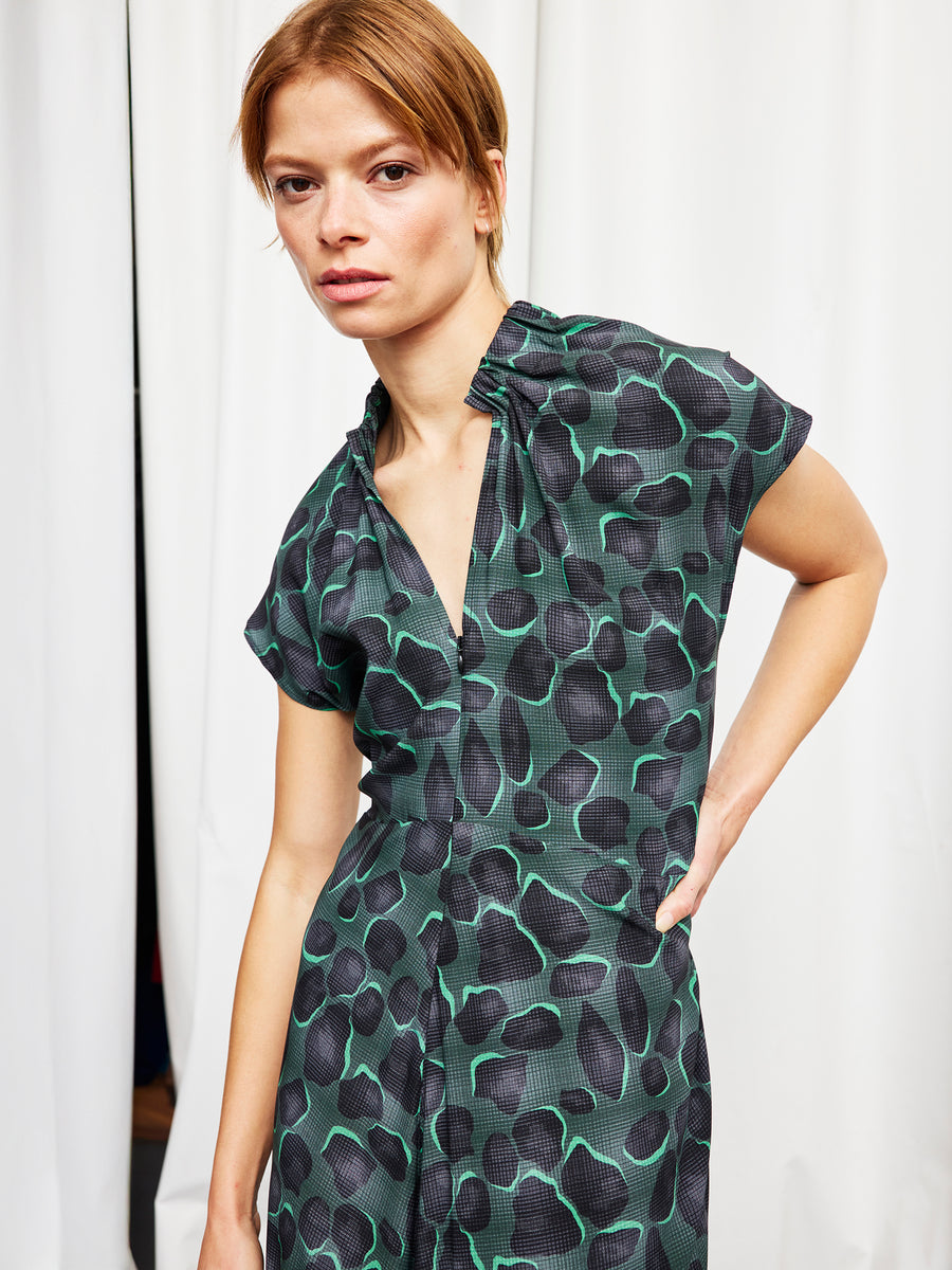 A woman with light brown hair wears the Zero + Maria Cornejo Long Ruched Stella Dress, featuring a black and green abstract pattern, and stands in front of white curtains looking at the camera with a neutral expression.