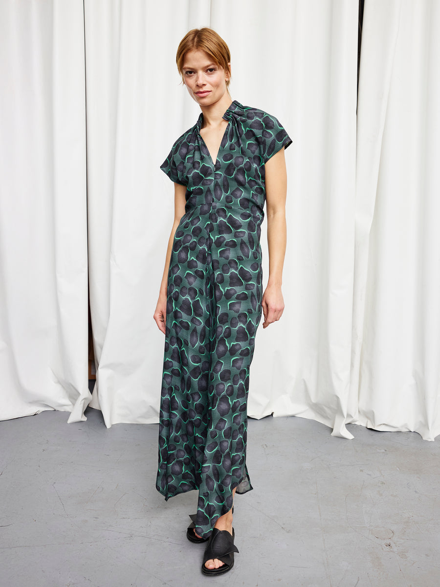 A person stands before white curtains wearing the Long Ruched Stella Dress by Zero + Maria Cornejo—full-length with a green and black circular pattern—paired with black open-toe shoes, short hair, and a neutral expression.