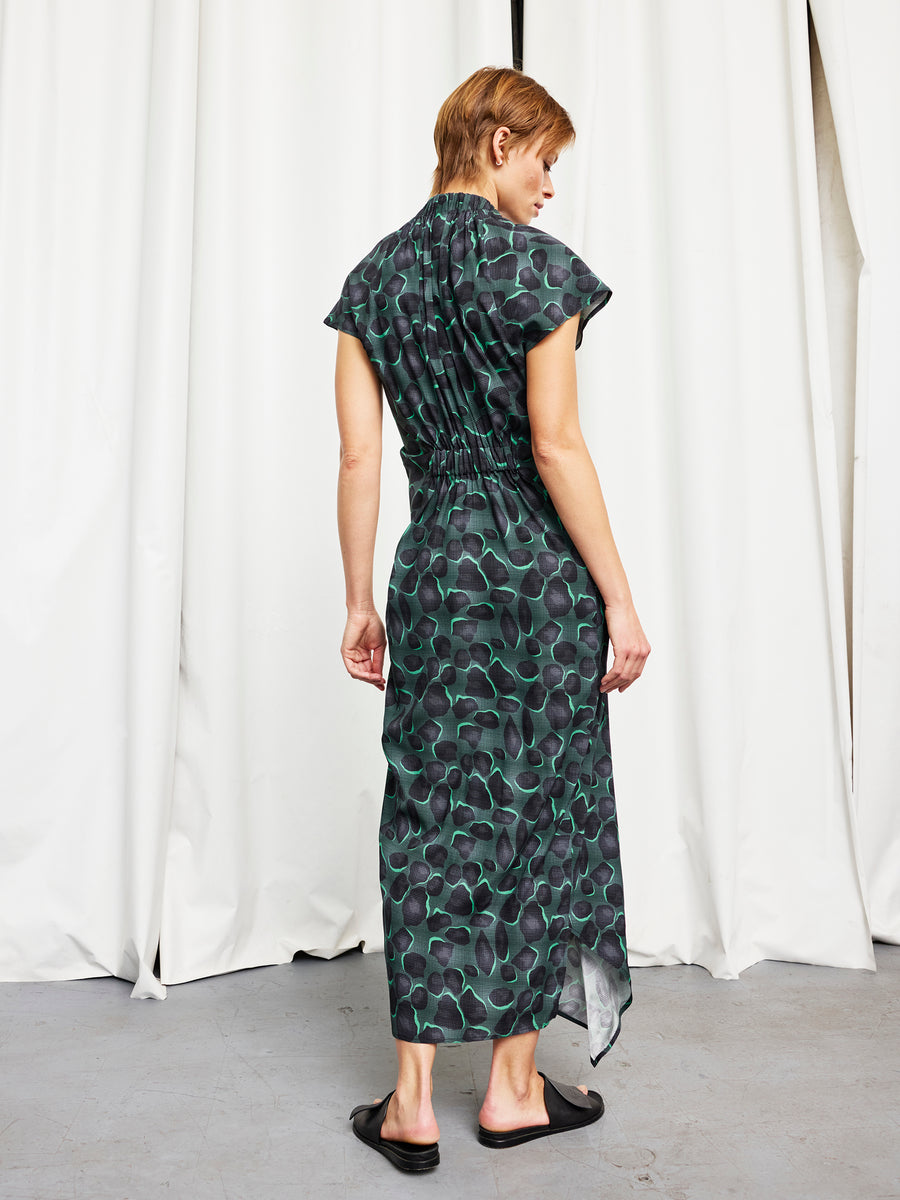 A person faces away, wearing the Long Ruched Stella Dress by Zero + Maria Cornejo— a dark green and black patterned, full-length, short-sleeved dress with a gathered waist—paired with black slip-on shoes against white curtains. Made in New York.