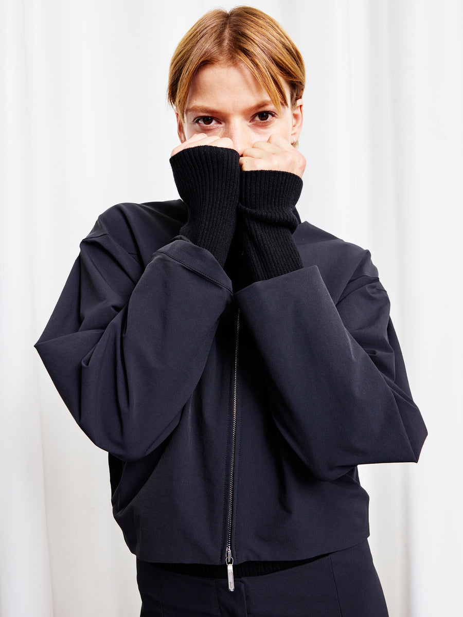 A person with short blonde hair stands against a white curtain, wearing the T Bomber by Zero + Maria Cornejo with black ribbed cuffs, holding their sleeves up to their face and looking directly at the camera.