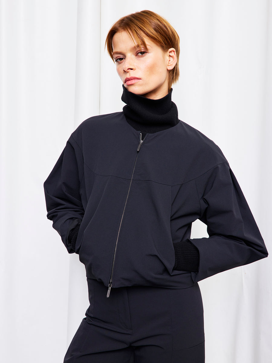A person with short light brown hair stands against a white curtain, hands in pockets, wearing Zero + Maria Cornejo’s black T Bomber jacket over a black turtleneck.