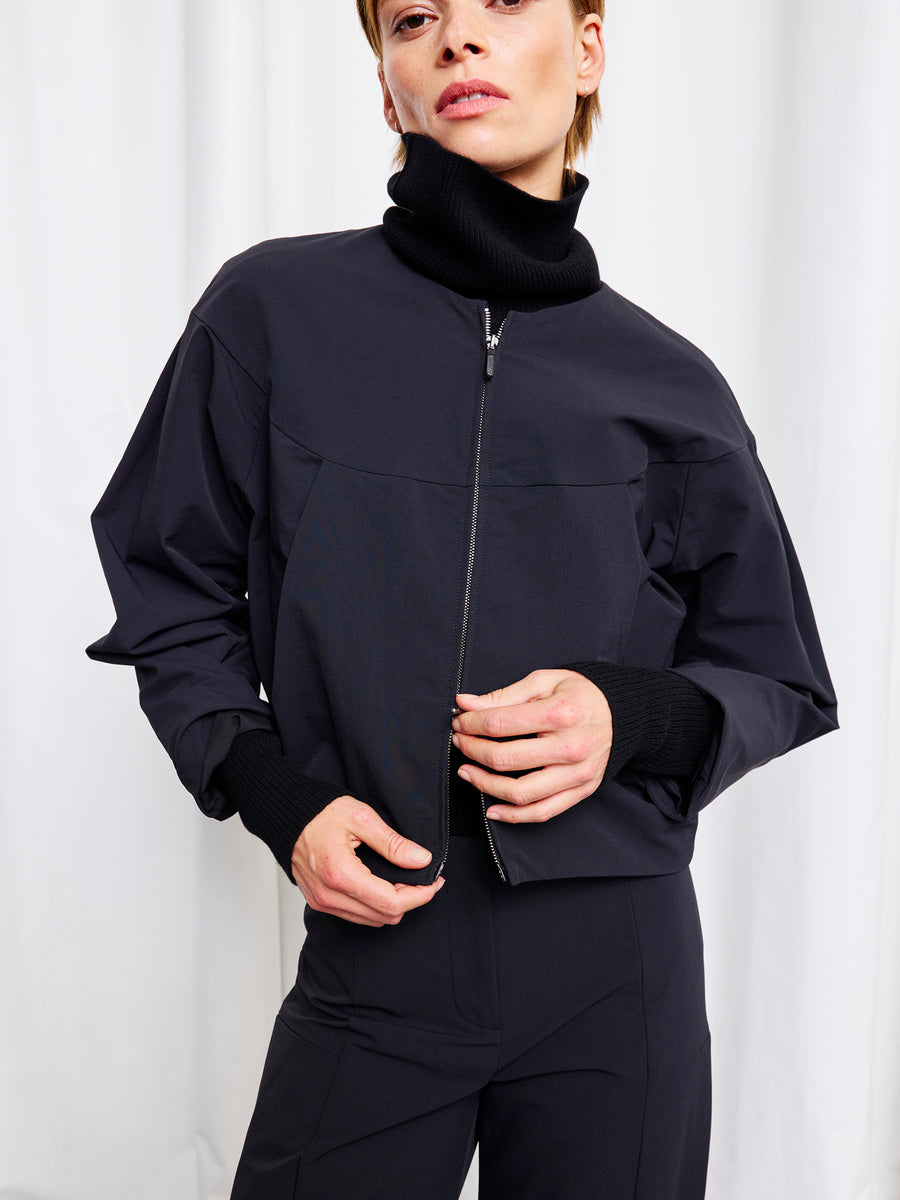 A person wearing the Zero + Maria Cornejo T Bomber, a black lightweight stretch jacket with a high collar, pairs it with black pants while standing in front of a white background. One hand adjusts the jacket, the other rests near the waist.