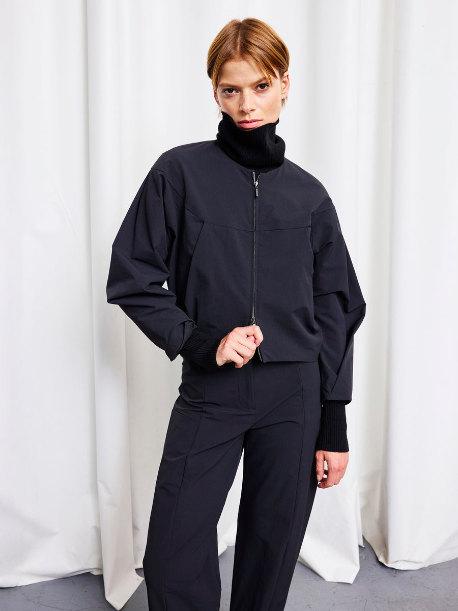 A person with short, light brown hair wears the Zero + Maria Cornejo T Bomber and matching pants, standing against white curtains and looking directly at the camera with a serious expression.