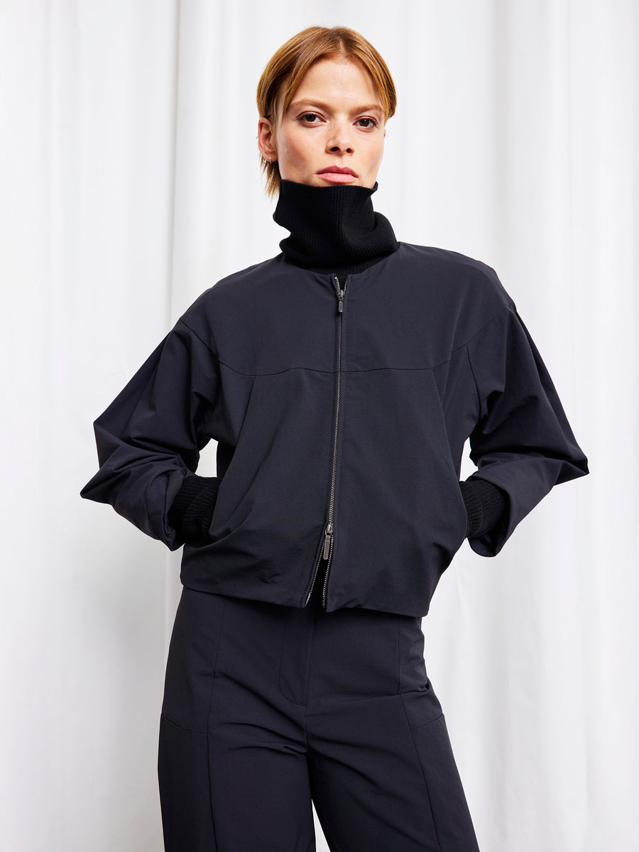 A person stands confidently with hands in pockets, modeling the black technical-stretch woven T Bomber by Zero + Maria Cornejo and matching turtleneck pants against white curtains. Made in New York.