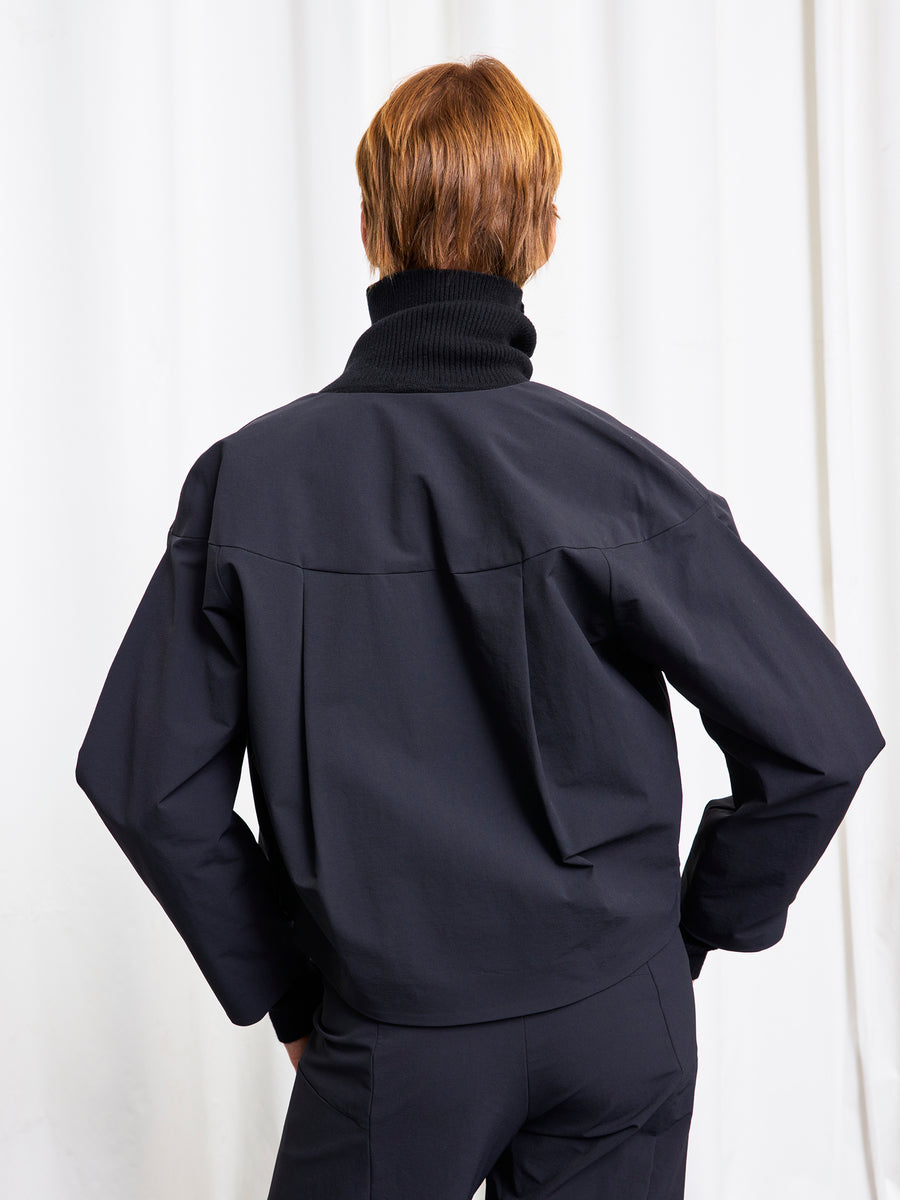 A person with short, light brown hair faces away, wearing a black turtleneck and Zero + Maria Cornejo T Bomber jacket and pants against a white background.