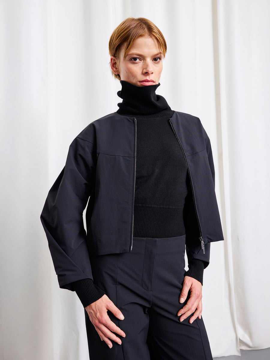 A person with short light brown hair wears a black turtleneck, black pants, and the T Bomber by Zero + Maria Cornejo, standing confidently before a white curtain. Made in New York.