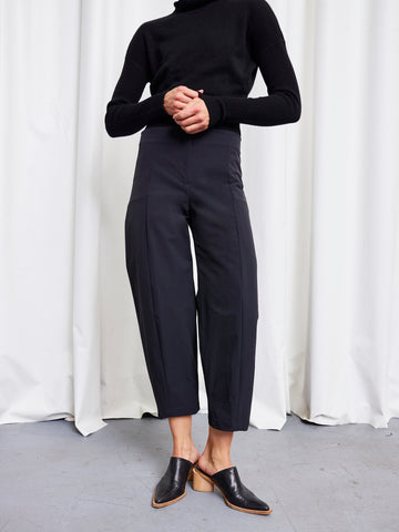 A person stands in front of white curtains, wearing a black turtleneck sweater and Zero + Maria Cornejo Margot Pants with black heeled mules. Their hands are clasped, and their head is out of frame.