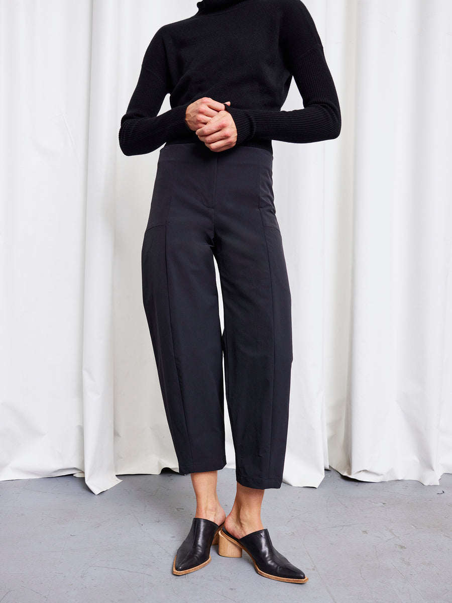 A person stands in front of white curtains, wearing a black turtleneck sweater and Zero + Maria Cornejo Margot Pants with black heeled mules. Their hands are clasped, and their head is out of frame.