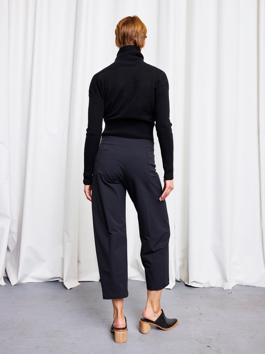 A person stands facing away from the camera, wearing a black turtleneck sweater, Zero + Maria Cornejo Margot Pant, and black open-back heeled shoes. White curtains hang in the background.