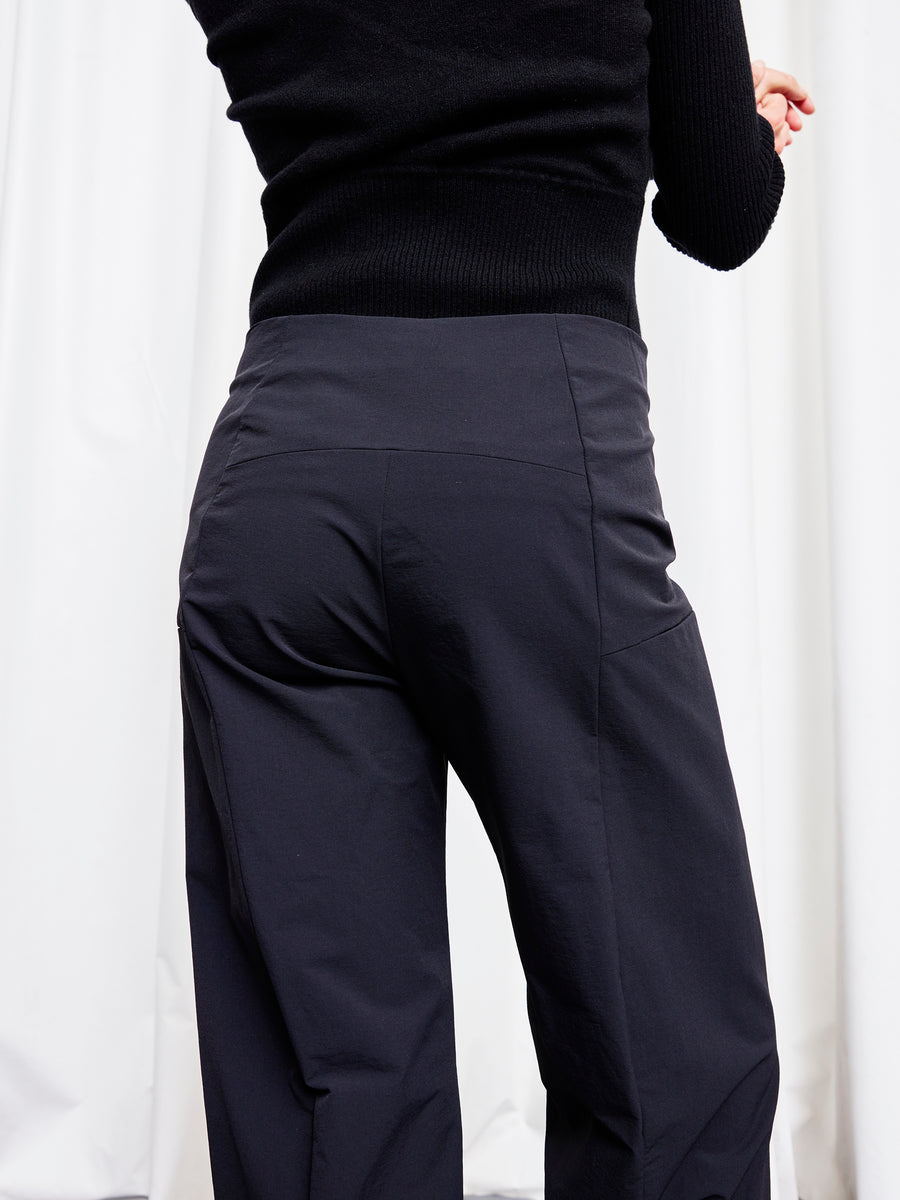 A person stands with their back to the camera, showcasing the fit and fabric of the Margot Pant by Zero + Maria Cornejo—a dark, wide-leg cropped style—paired with a black sweater in front of white curtains.