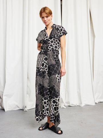 A person stands indoors against white curtains, wearing the Zero + Maria Cornejo Long Ruched Stella Dress— a full-length black and white patterned silk charmeuse dress with short sleeves, made in New York—paired with black slide sandals.