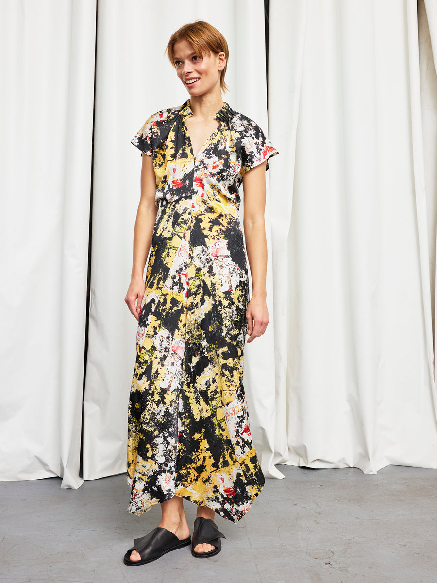 A person stands before white curtains, smiling and looking to the side, wearing the Long Ruched Stella Dress by Zero + Maria Cornejo in a colorful abstract print, paired with black slide sandals.