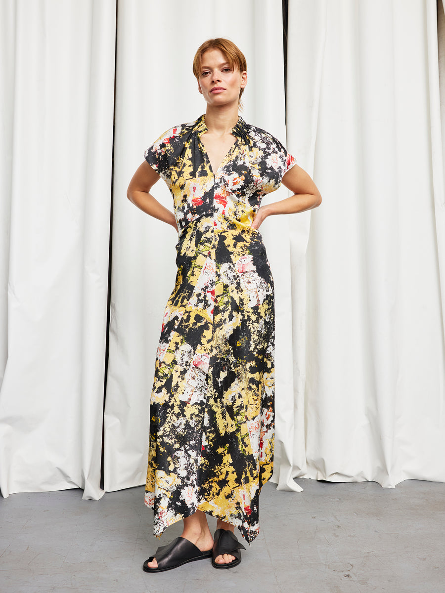 A person stands with hands on hips, wearing the Long Ruched Stella Dress by Zero + Maria Cornejo in a full-length floral pattern, paired with black slide sandals. White curtains hang in the background. Made in New York.