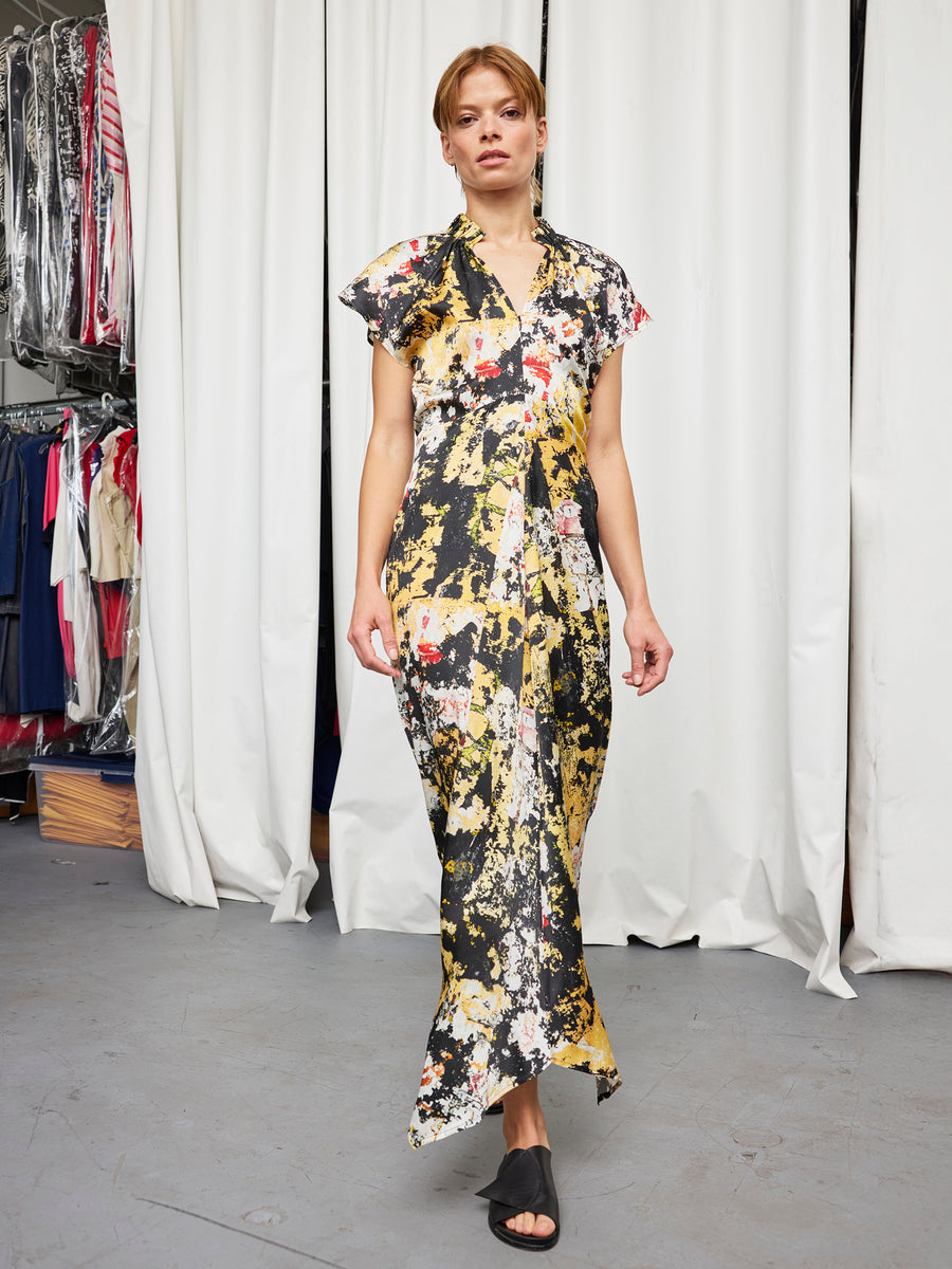 A woman with brown hair tied back wears the Zero + Maria Cornejo Long Ruched Stella Dress, featuring short sleeves and an abstract yellow, black, red, and white print. She stands indoors before white curtains and clothing racks in black open-toe shoes.