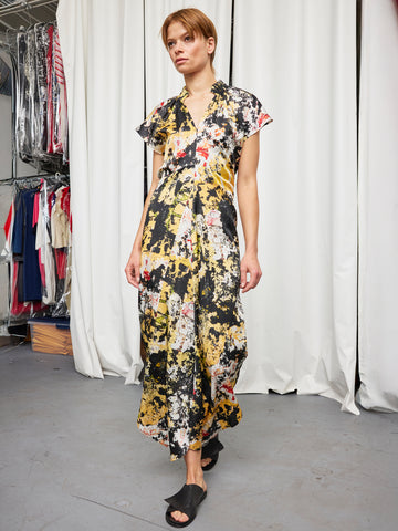 A person with short hair wears the Long Ruched Stella Dress by Zero + Maria Cornejo, featuring a loose, colorful abstract pattern, with black slide sandals in a room with white curtains and clothing racks.
