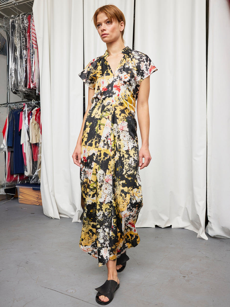 A person with short hair wears the Long Ruched Stella Dress by Zero + Maria Cornejo, featuring a loose, colorful abstract pattern, with black slide sandals in a room with white curtains and clothing racks.