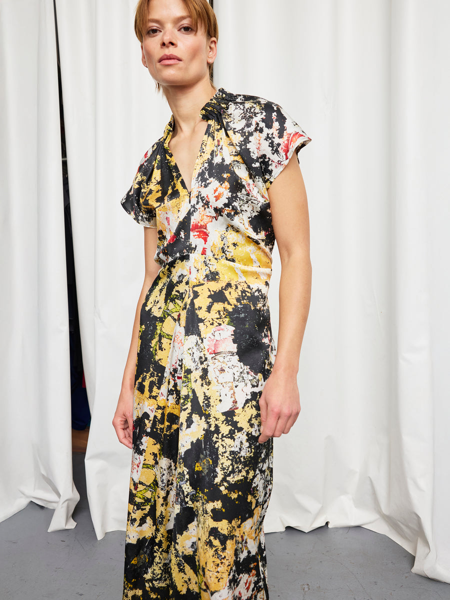 A person stands in front of white curtains wearing the Zero + Maria Cornejo Long Ruched Stella Dress, a short-sleeved, V-neck silk charmeuse featuring an abstract yellow, black, and white splatter pattern.