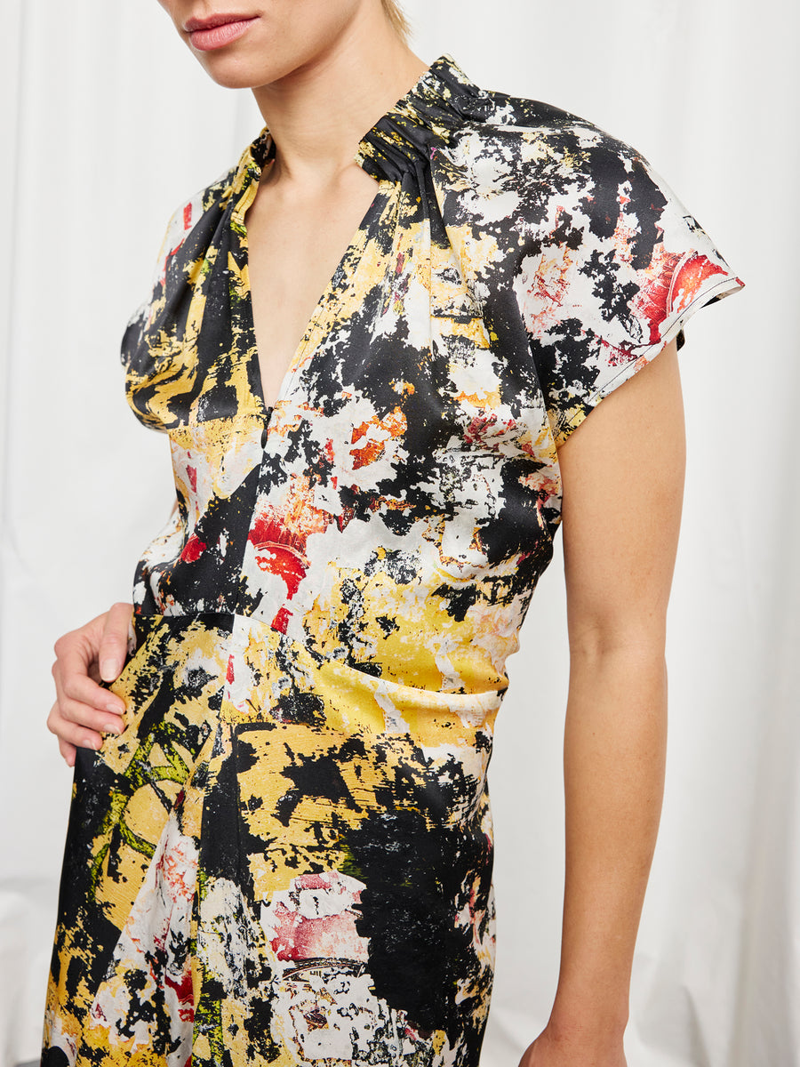 A person wearing the Zero + Maria Cornejo Long Ruched Stella Dress, a short-sleeve silk charmeuse with a yellow, black, red, and white abstract paint splatter pattern, stands against a white background. Only their upper body is visible.