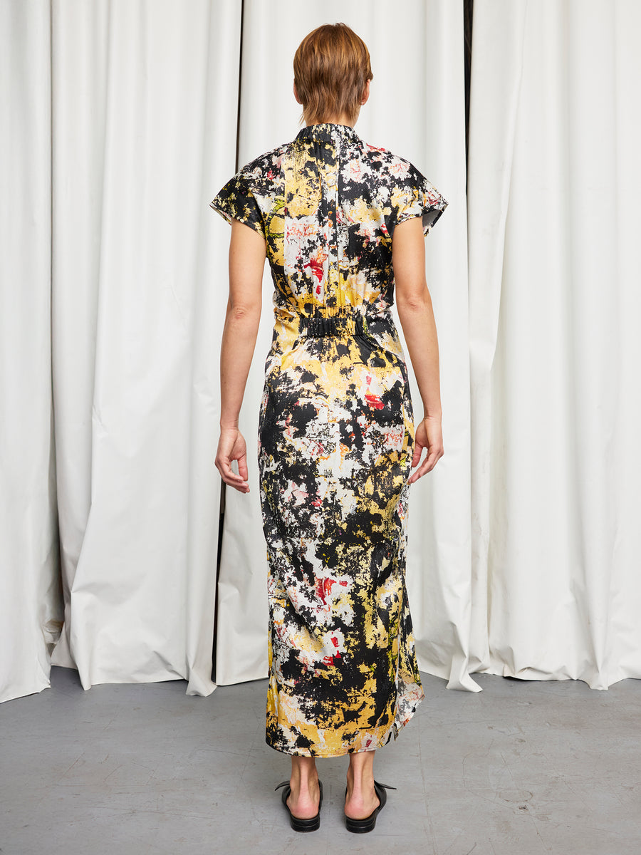 A person with short brown hair stands facing away, wearing the Zero + Maria Cornejo Long Ruched Stella Dress in a black, yellow, white, and red abstract print with black sandals. White curtains are in the background.