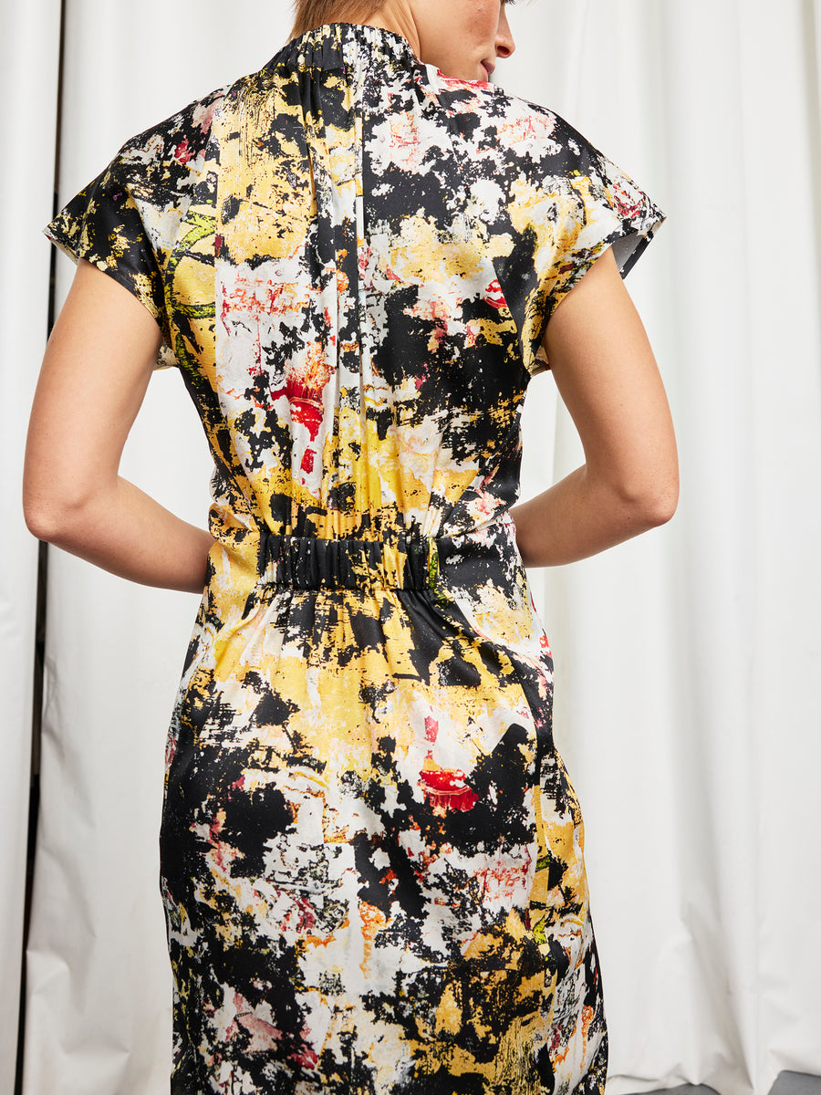 A person stands with their back to the camera, wearing the Zero + Maria Cornejo Long Ruched Stella Dress in a short-sleeved silk charmeuse with an abstract black, yellow, white, and red splatter pattern. White curtains are in the background.