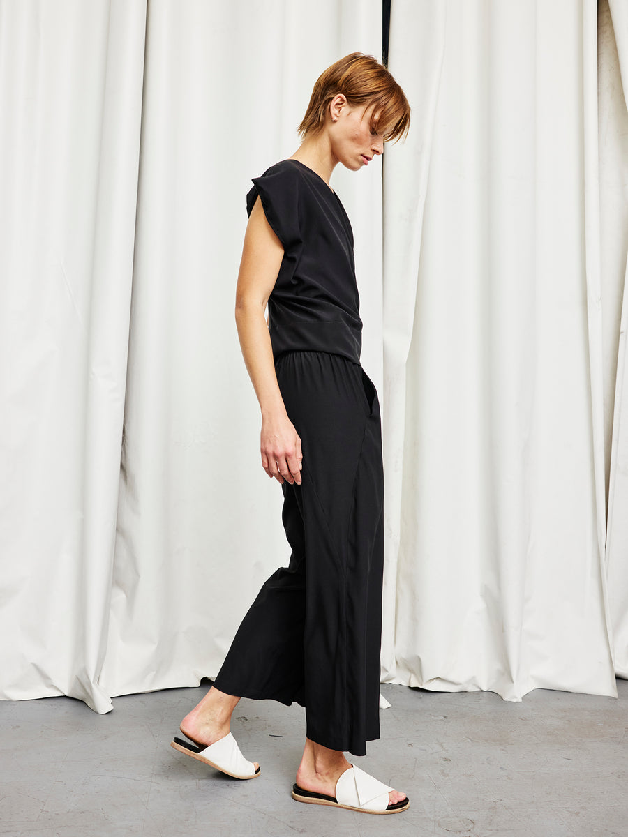 A person with short hair wears a sleeveless black jumpsuit and white slide sandals, standing in profile against white curtains and a gray floor—an effortlessly chic look, completed with the Ido Pant by Zero + Maria Cornejo.