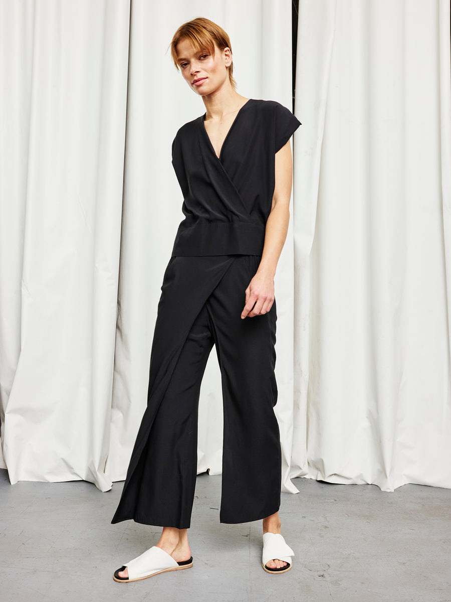 A person stands before white curtains wearing the Zero + Maria Cornejo Ido Pant in black with wide legs, paired with white slide sandals, and looks at the camera with a slight smile.