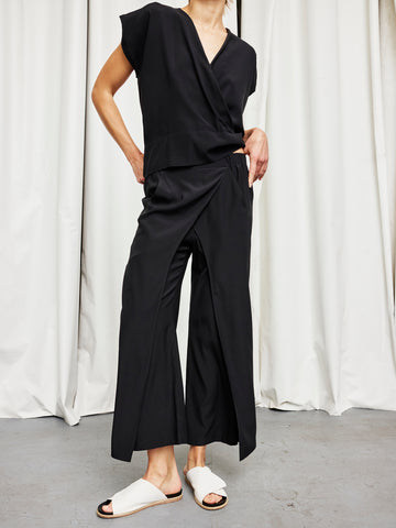 A person stands in front of white curtains wearing a loose black wrap top and the Ido Pant in black by Zero + Maria Cornejo, paired with white slide sandals. Their face is out of frame.