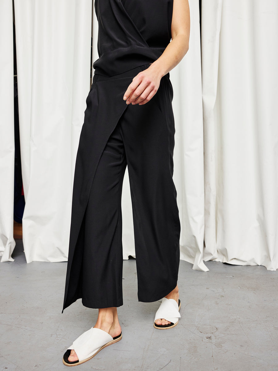 A person wears the Zero + Maria Cornejo Ido Pant in black silk charmeuse with a sleeveless black top and white open-toed slide sandals, standing on a gray floor before white curtains. Their face is not visible.