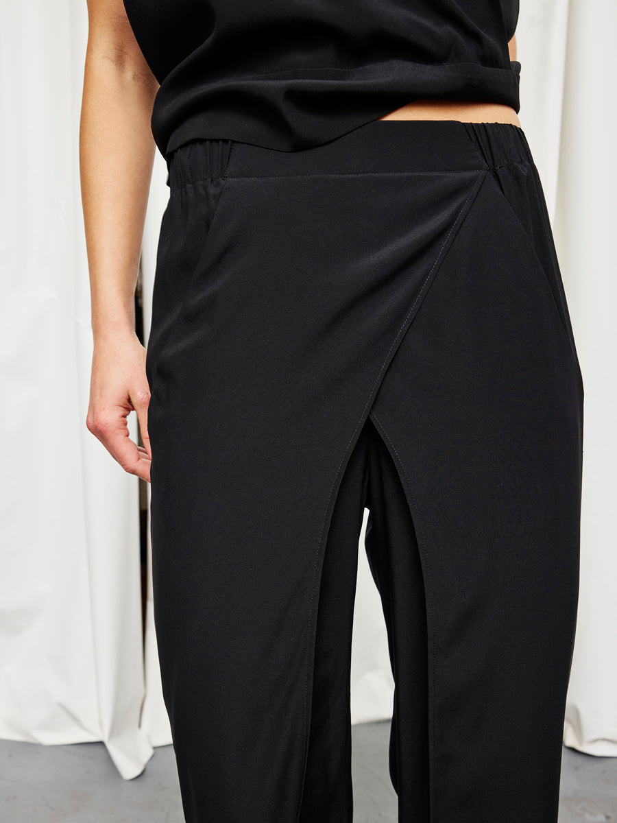 A person models the Zero + Maria Cornejo Ido Pant—black stretch silk pants featuring a front overlapping wrap design—shown from the lower torso to upper legs, posed against a white curtain backdrop.