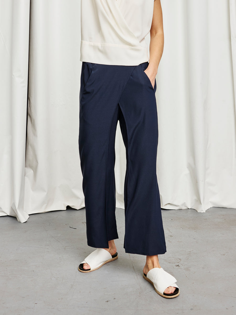 A person stands with one hand in their pocket, wearing the Ido Pant by Zero + Maria Cornejo—navy, wide-leg, stretch silk—paired with a sleeveless white top and white open-toe slide sandals. White curtains and a gray floor set the scene. Made in New York.